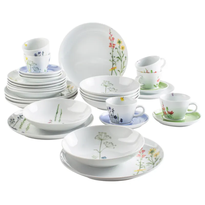 Eine Sammlung von Geschirr, bestehend aus weißen Tellern, Schalen und Tassen mit bunten, floralen Motiven. Im Vordergrund sind mehrere flache Teller, Schalen und unterschiedliche Tassen mit passenden Untertassen angeordnet. Im Hintergrund sind weitere Geschirrteile gestapelt. Die floralen Designs verleihen dem Geschirr eine fröhliche und lebendige Ausstrahlung.