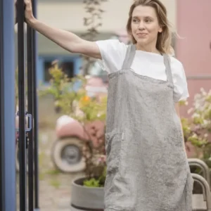 Woman standing in a grey striped linen cross-back apron, resting her arm on an open door frame outdoors Epic Linen premiuim linen