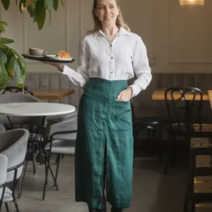 Woman standing in a café wearing a dark green linen waist apron over a white button-down shirt, holding a serving tray with a coffee cup and pastry.